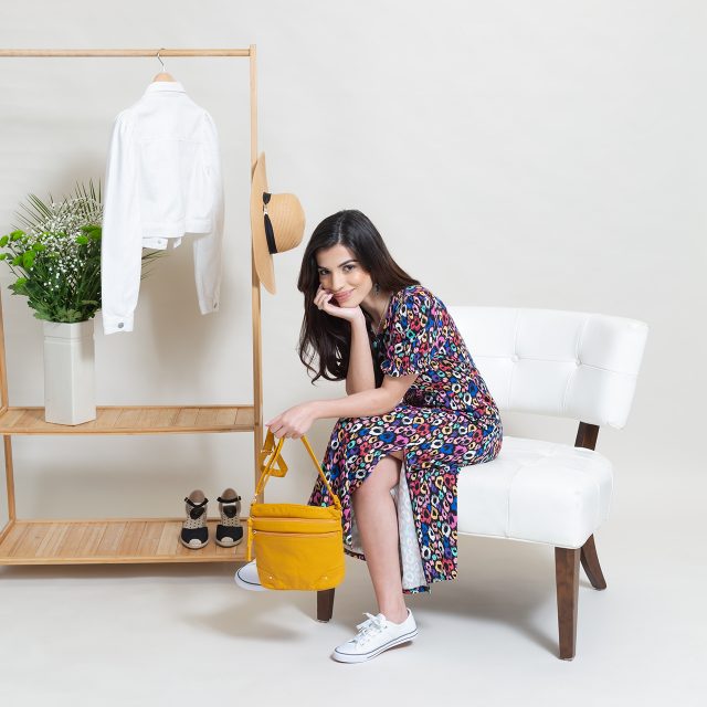 A model eCommerce shot showing a model sat down in front of a clothes rail. She's holding a yellow bag and wearing a bright, colourful dress.