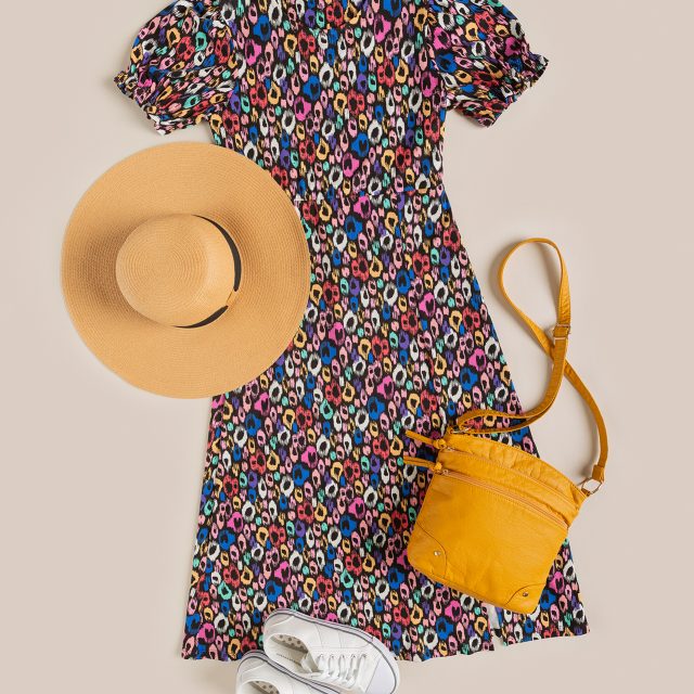 An eCommerce flatlay shot, taken from above. It shows the same colourful dress from earlier arranged with a yellow bag, white trainers, and a beige sun hat.